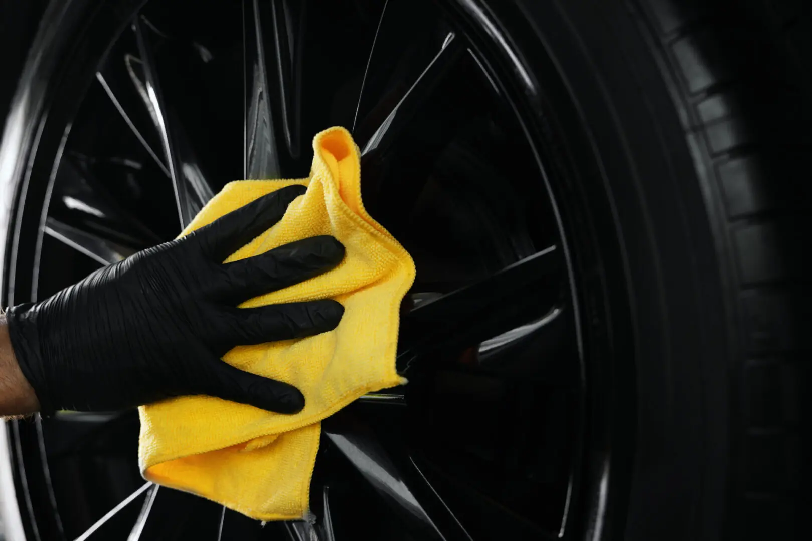 A hand in a black glove cleans a black car wheel with a yellow cloth.