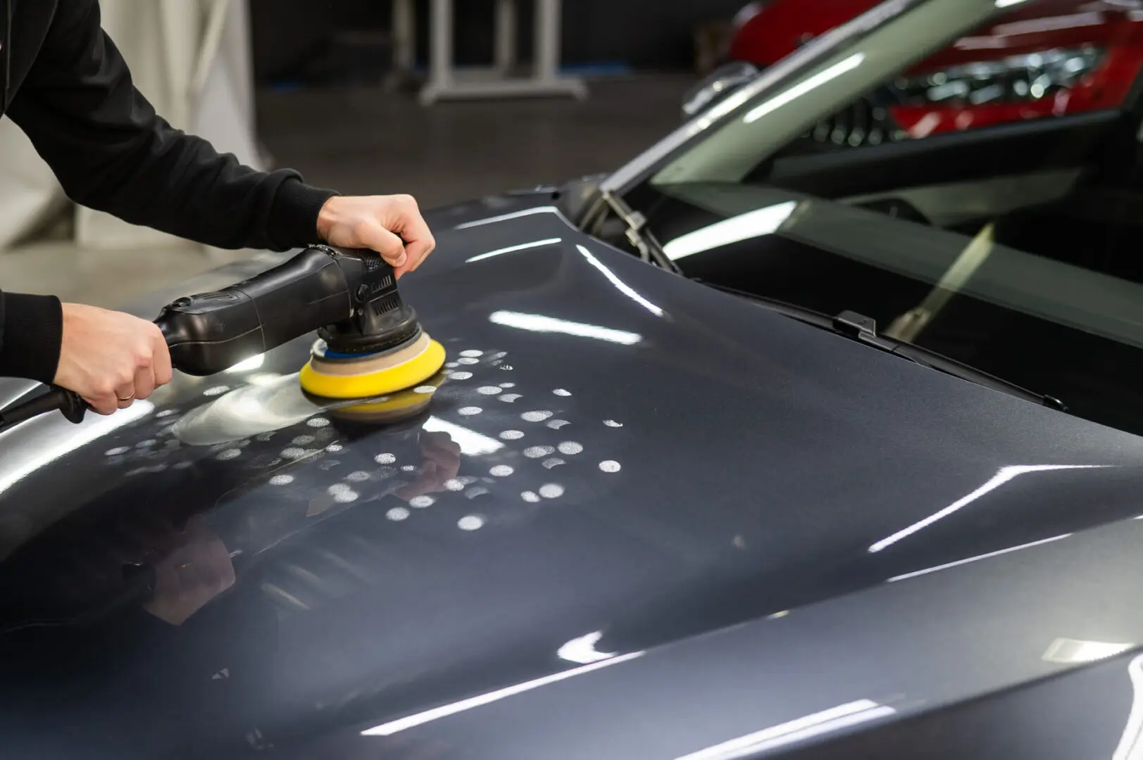 Person buffing a car hood with an electric polisher, removing blemishes from the dark-colored surface in a garage setting.