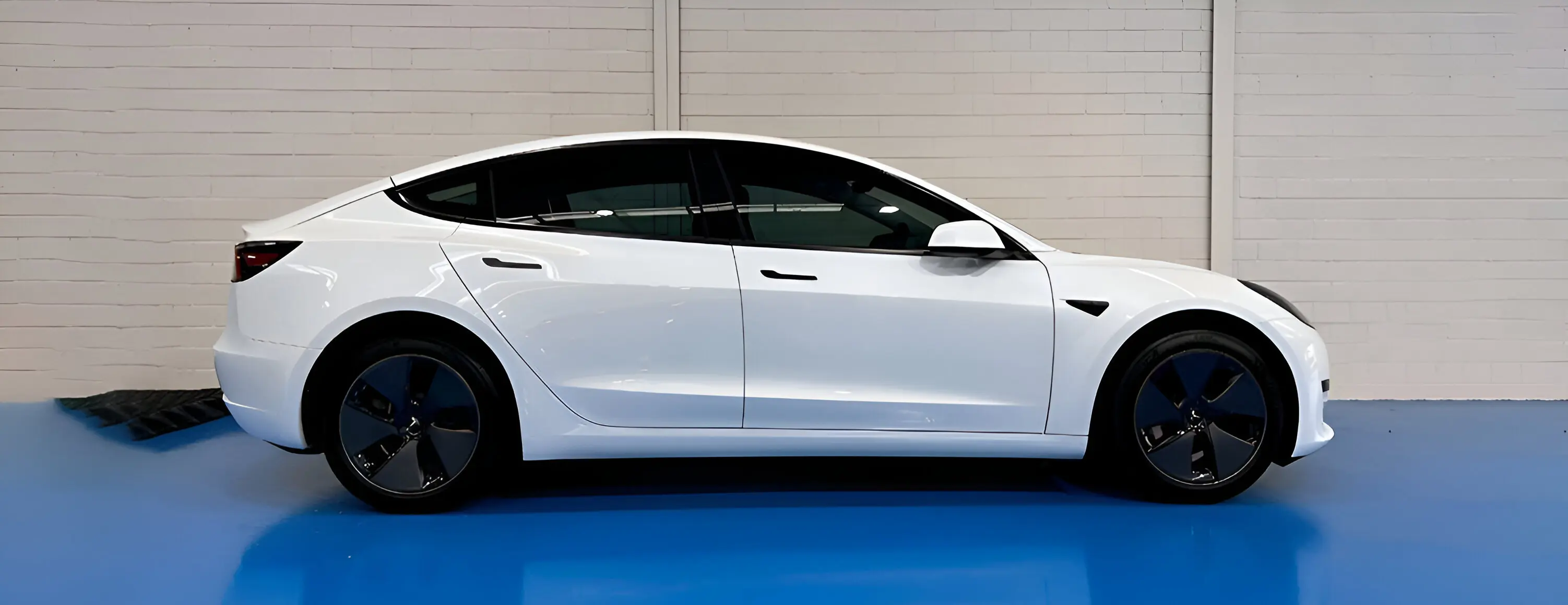 White electric car parked indoors on a blue floor, viewed from the side against a light brick wall.